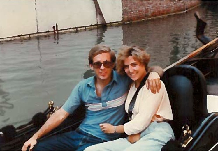 A man and a woman sit together in a gondola. The man's arm is around the woman's shoulder. Both are smiling.