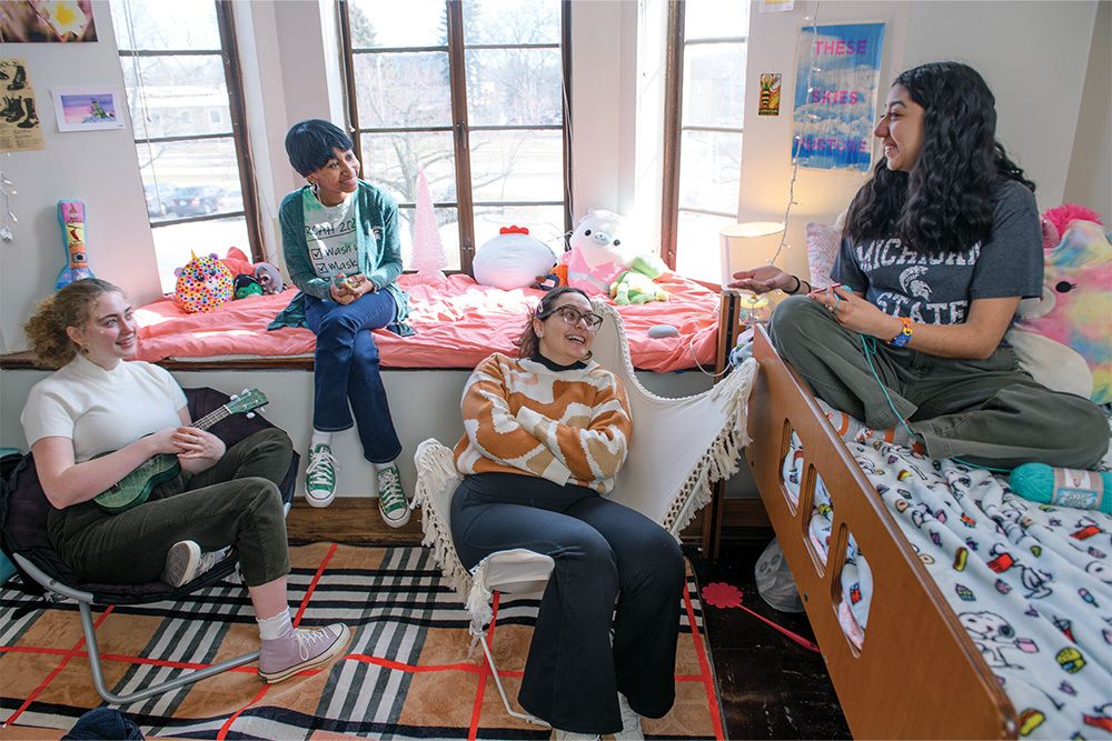 Four smiling students leisure around a dorm room conversing with one another. Two students are sitting in chairs while the third sits in a window alcove facing another in a dorm bed.