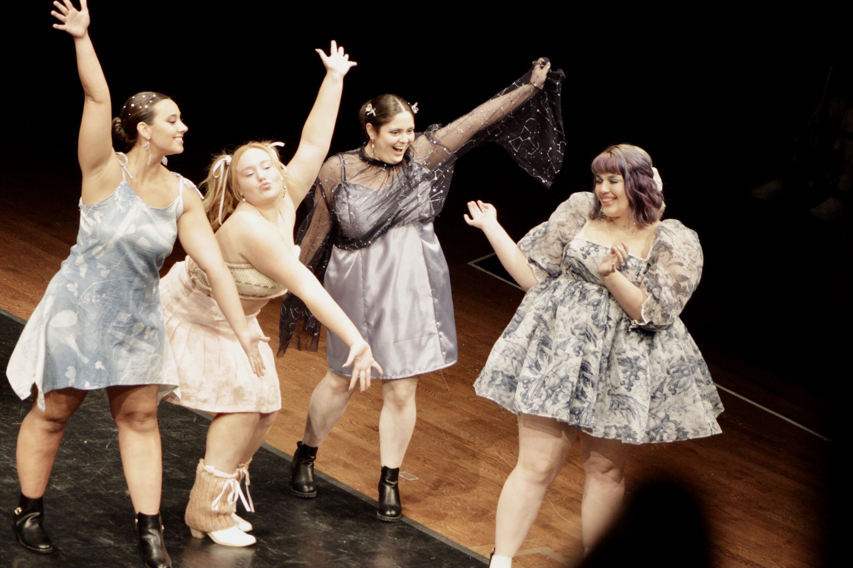 On a stage, four people in dresses with arms out in excitement.