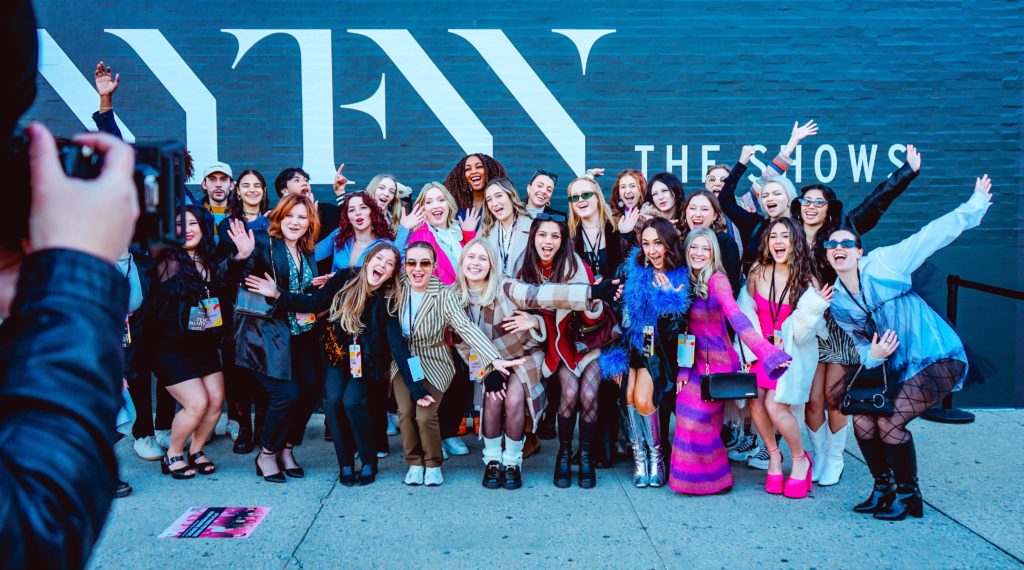 A group of 30 smiling students wearing elaborate outfits stand posing for the camera against a blue wall that reads "NYFW The Shows."