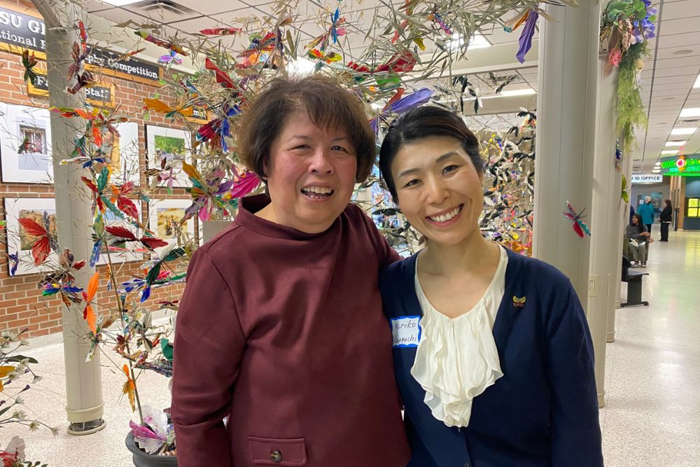 A picture of two women side by side. The woman on the left has short brown hair and wears a red shirt; the woman on the left has black hair pulled back and wears a white and blue shirt. The are posing before an art exhibit.