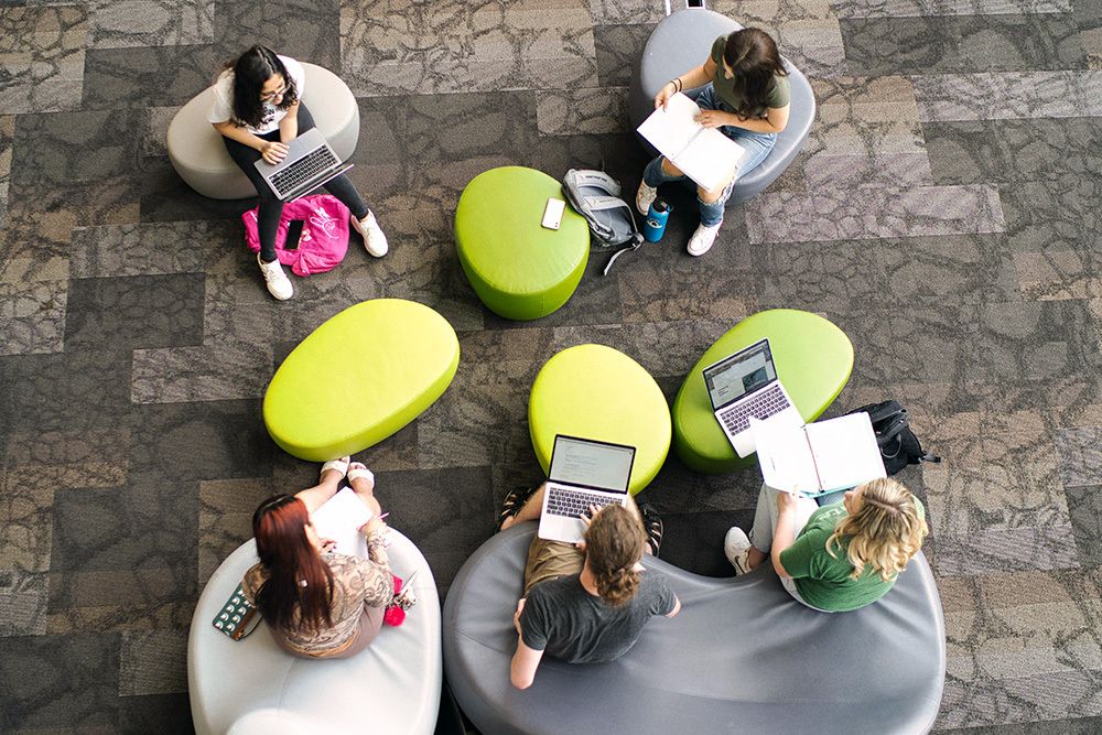 A bird's-eye view of a group of students sitting around green stools.