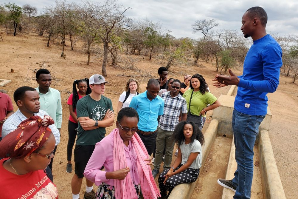 A man wearing a blue long sleeve shirt and jeans stands over of a group of people in the African plains with small trees in the background.