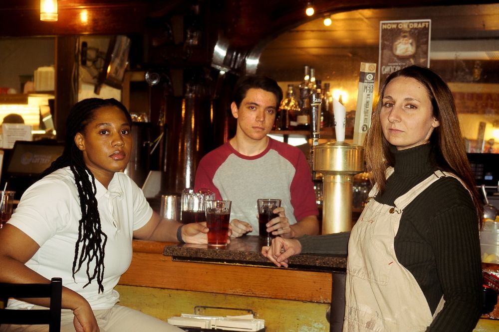 A man stands behind a counter while two women stand in front of it in a warmly lit bar. Their faces are expressionless as they gaze at the camera.