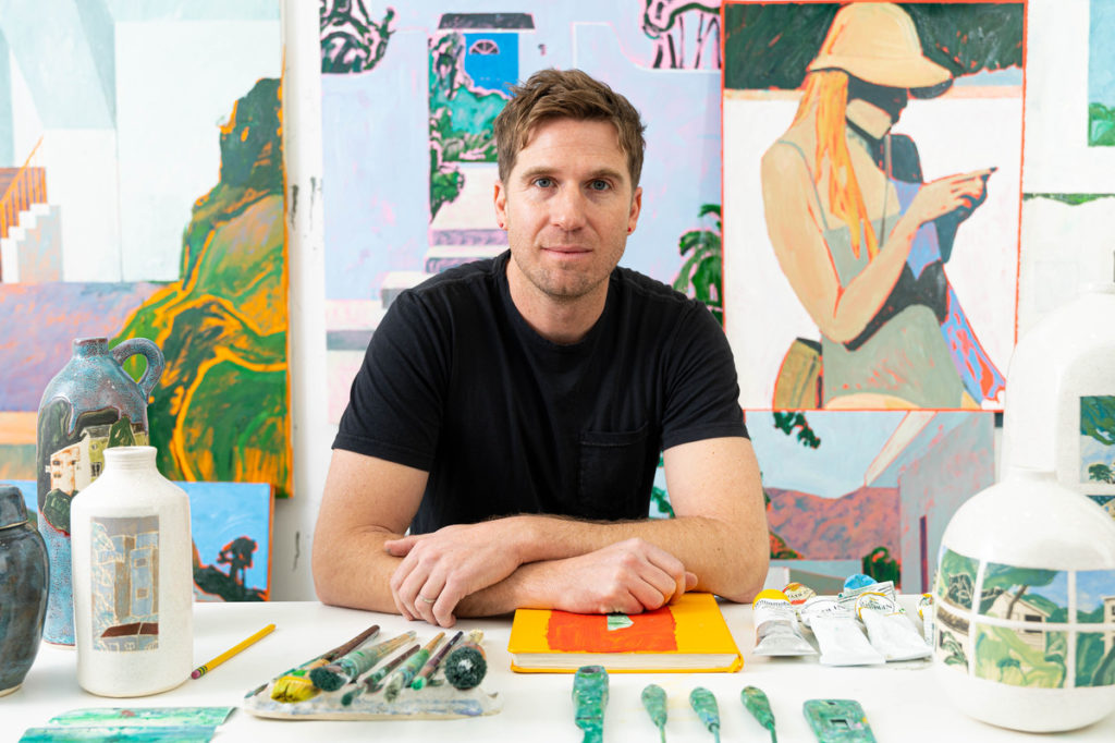 A man sits behind a desk with paintbrushes and painted sculptures with paintings in the background,