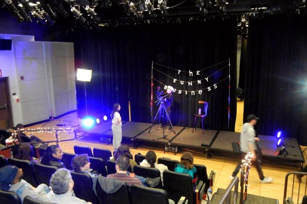 Photo showing a stage with the words "The Funny Business" spread across the black curtains. 