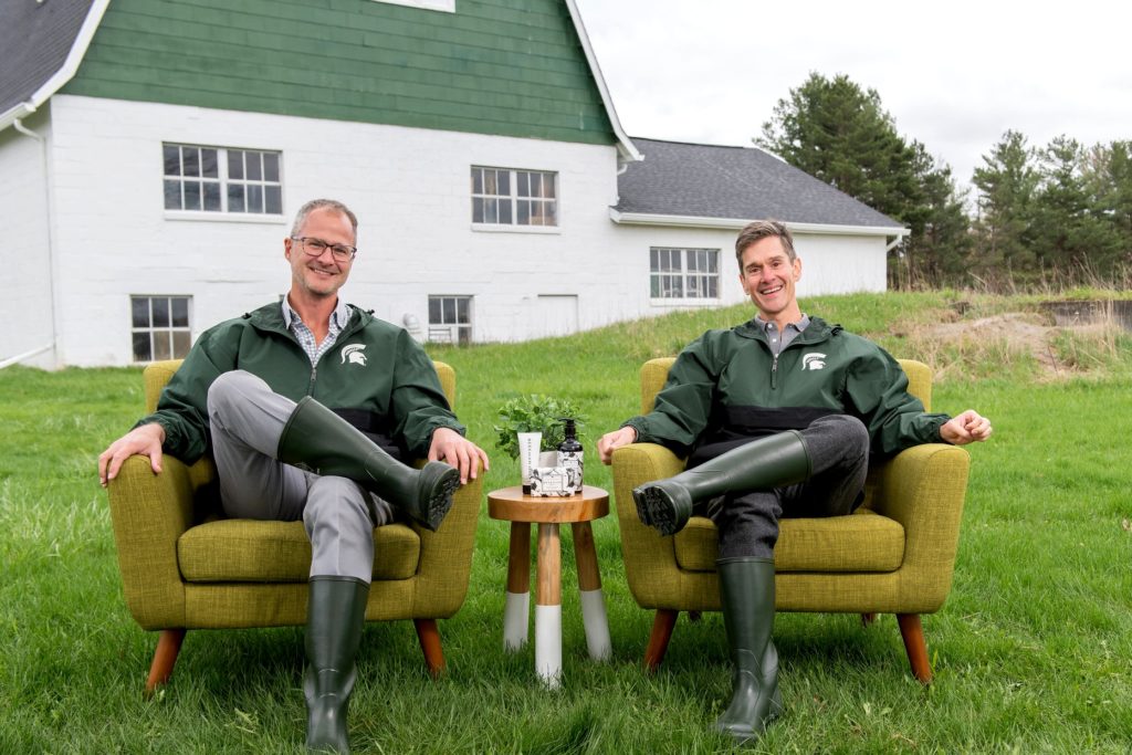 Josh Kilmer-Purcell and his husband Dr. Brent Ridge sitting on chairs in front of a barn next to their Beekman 1802 skincare products.