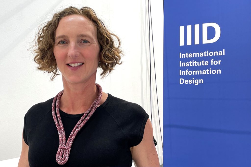 A woman with curly hair, Kelly Salchow MacArthur, stands in front of sign that says, "IID International Institute for Information Design."