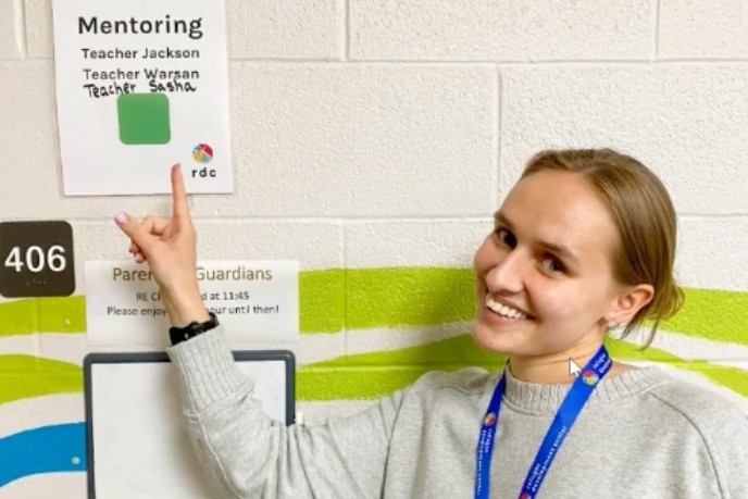Photo of a woman (Sasha Beloglazova) who is pointing at a sign that says "Mentoring" at the top and Sasha's name is listed below that along with two other names. 