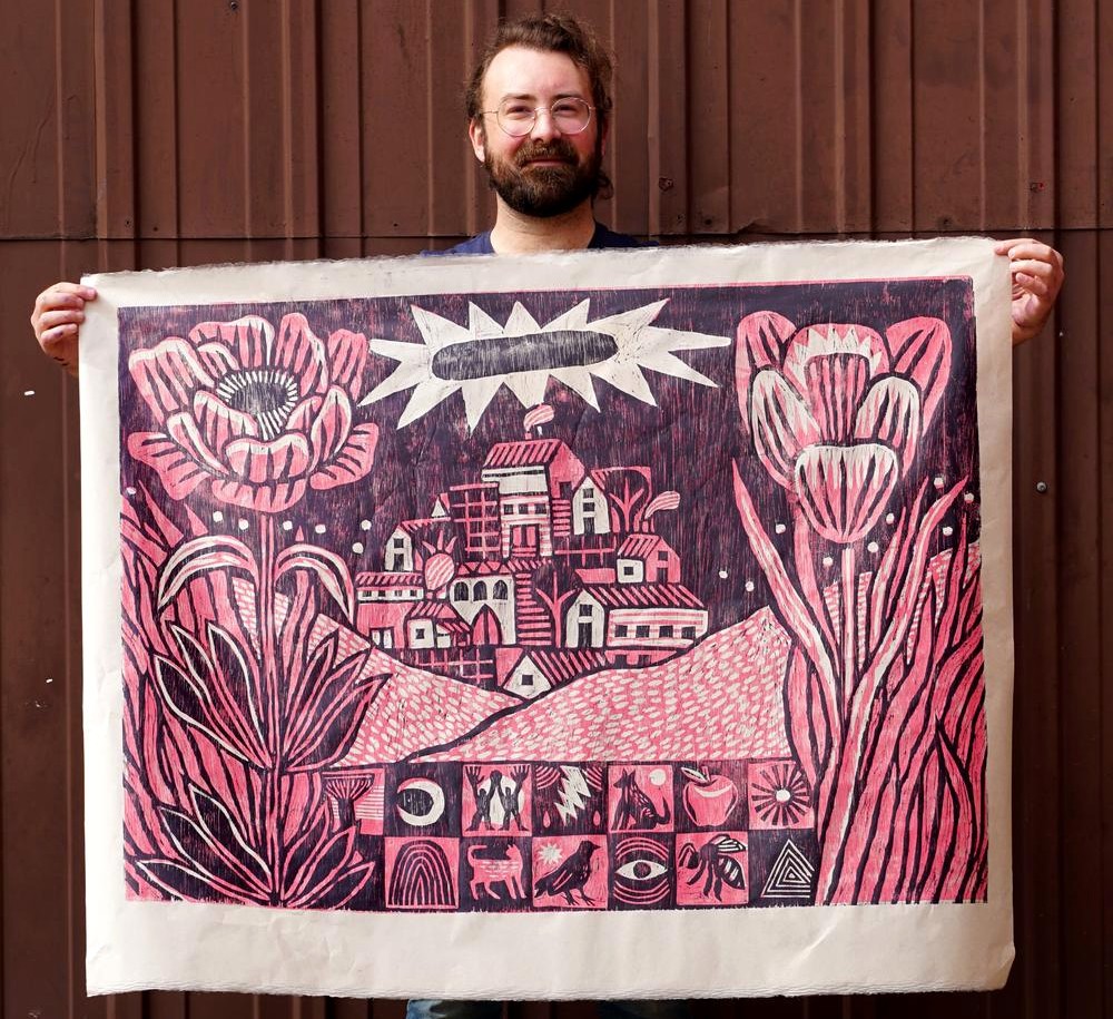 Photo of Trevor Grabill holding a large, original print of a town in the distance, nestled between a field and two large flowers. 