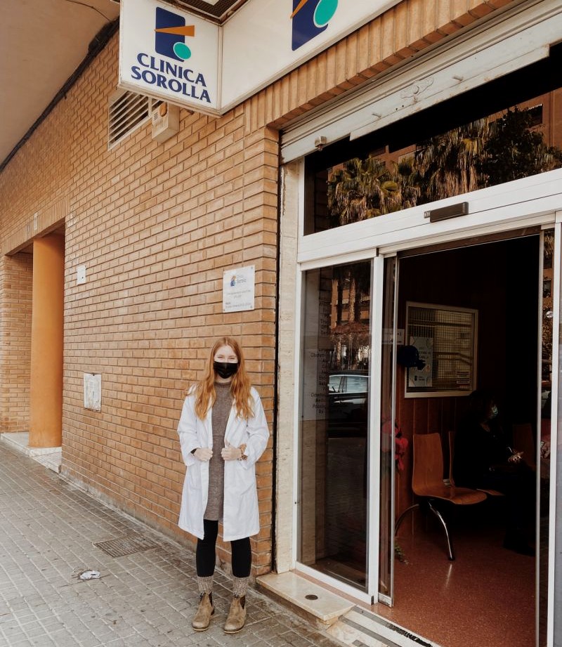Woman with long hair wearing a black face mask and a white lab coat standing outside the front doors of a clinic with a sign that says "Clinica Sorolla" overhead. 