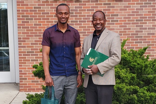 Two men standing in front of brick wall. One of the men is holding a green folder with the MSU logo on it. 