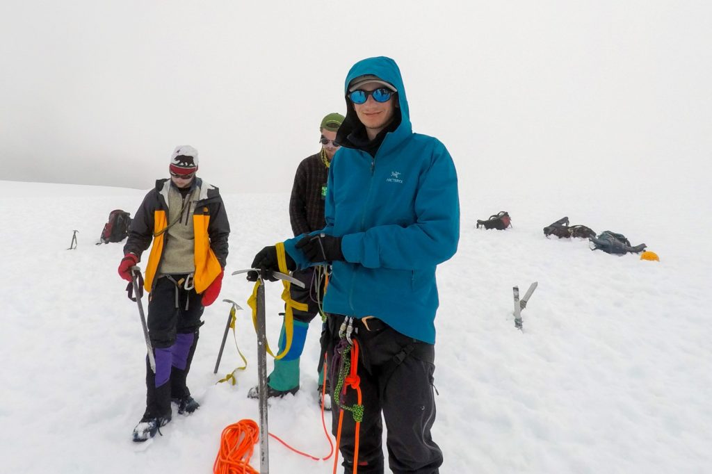 A picture of a man standing in front of a group on people on a glacier. He is wearing a bright blue coat and carrying an ice axe.