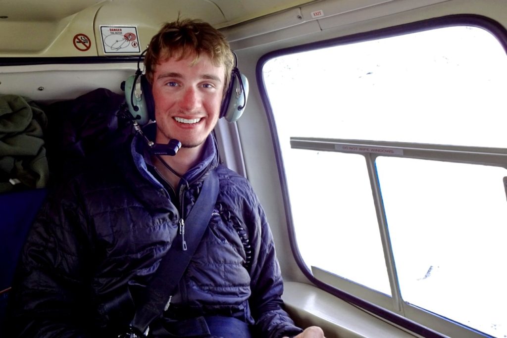 A picture of a man in a helicopter with a headset on. He wears a black jacket. 