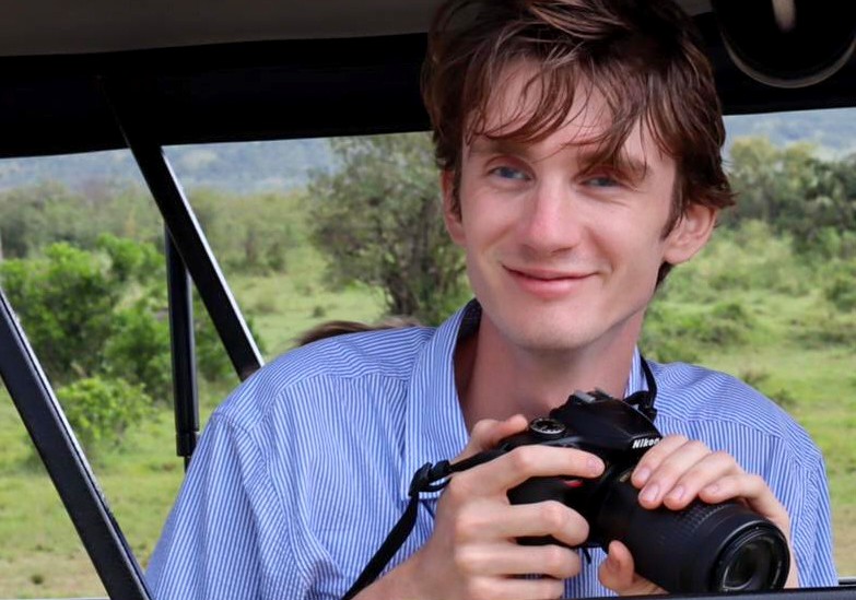 A picture of a man with a camera in his hands. He has brown hair and a while-and-blue striped shirt. 