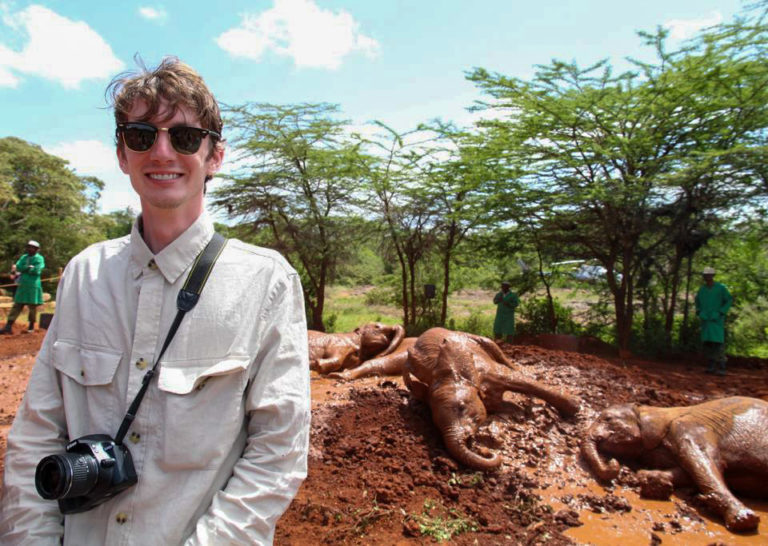 A picture of a man wearing a white shirt with a camera and sunglasses. Behind him are elephants.