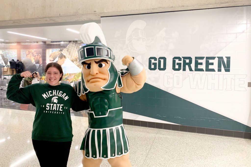 Photo of person standing with a spartan mascot in front of wall that says “Go green, go white.”