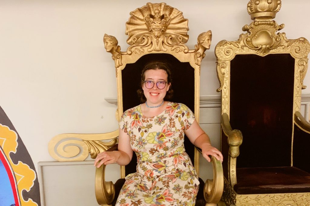 Photo of someone sitting down in a floral dress, in a chair that is gold and large.