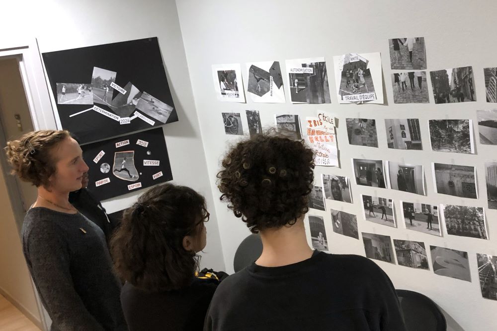 Three people look at an exhibit of photos and collages posted on a wall.