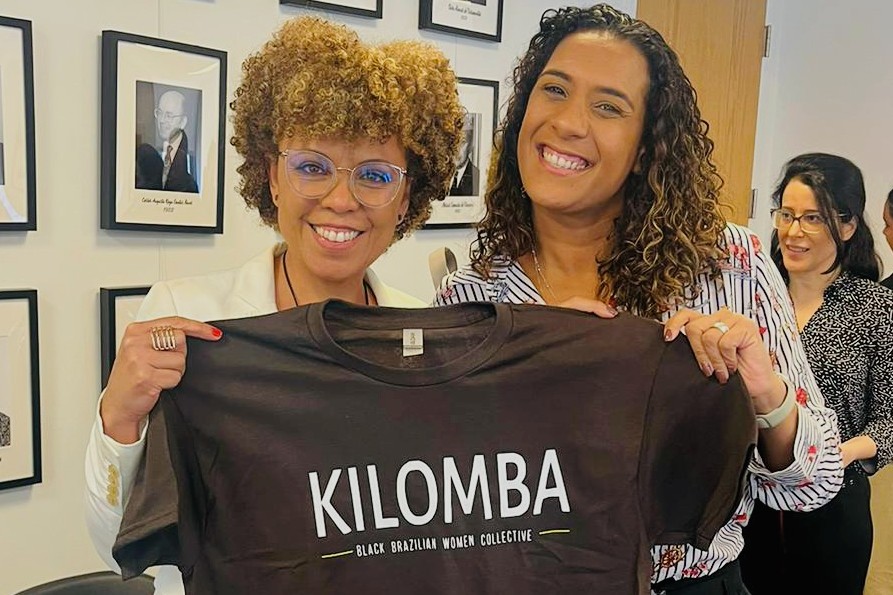 Two women standing together holding a T-shirt that says "Kilomba: Black Brazilian Women Collective:
