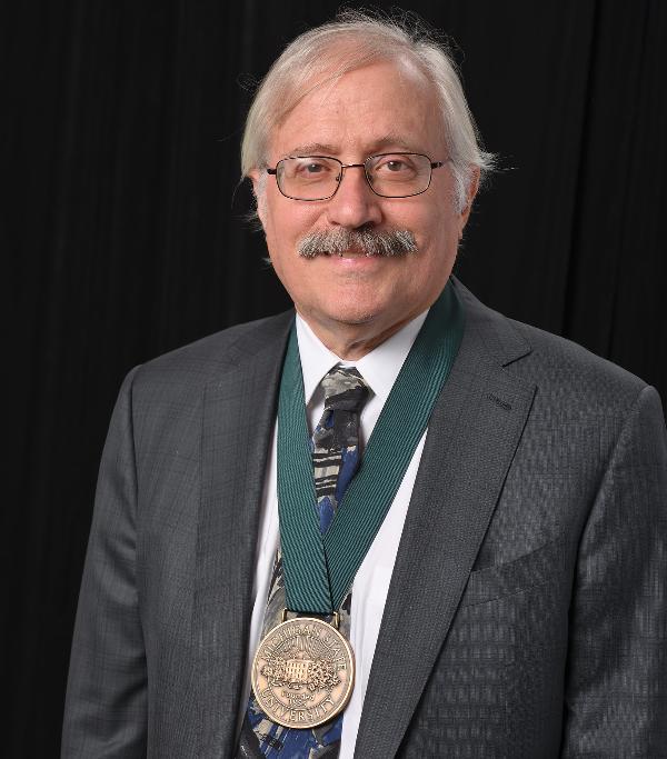 Man with gray hair and glasses with a medallion around his neck.