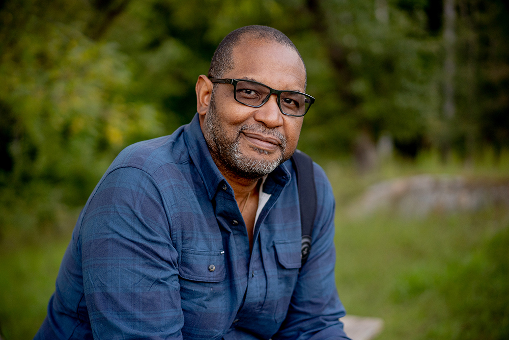 Photo in an outdoor setting of Dr. J. Drew Lanham - a Black man with black-rimmed glasses, short beard and moustache, shaved head with black stubble, and wearing a blue plaid button-up shirt. 