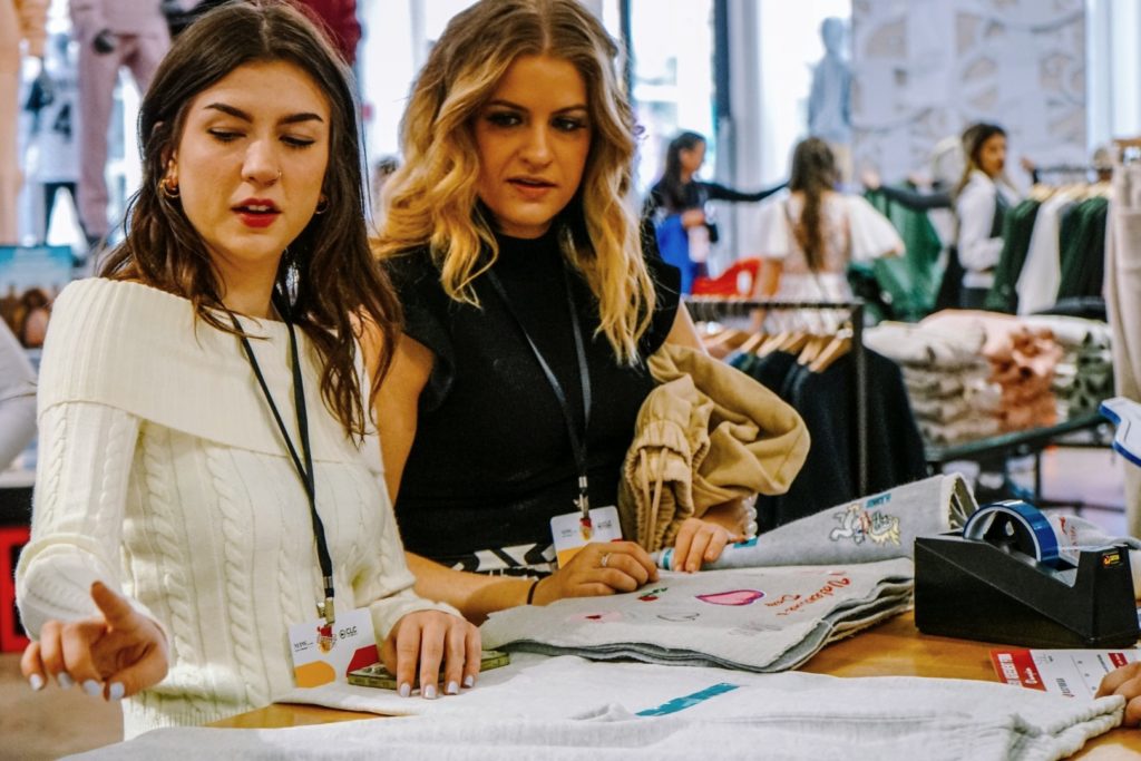 A picture of two people examining fashion material. The person on the left wears a white sweater while the person on the right wears a black top. 