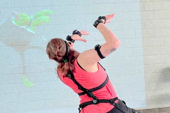Photo of woman in a strange position with technology strapped to her joints. In front of her a green animated character in following her same movement.