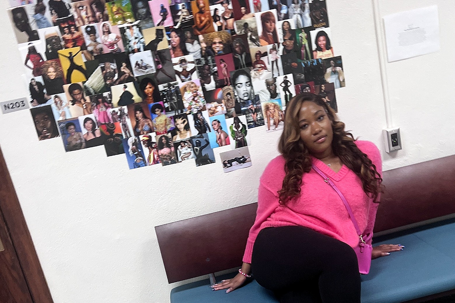Woman sitting on a couch looking at camera with many photos pasted on a white wall behind her.