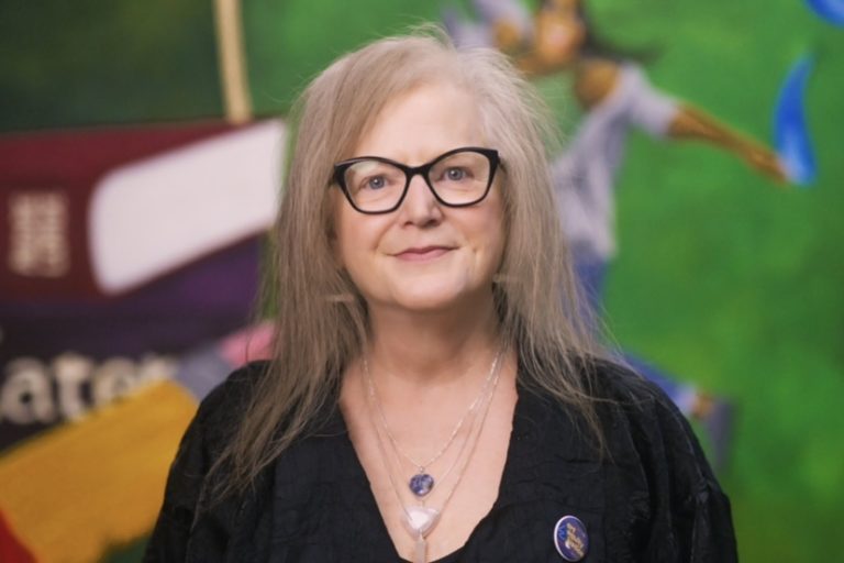 A woman in a black shirt with black glasses and necklace and blonde hair.