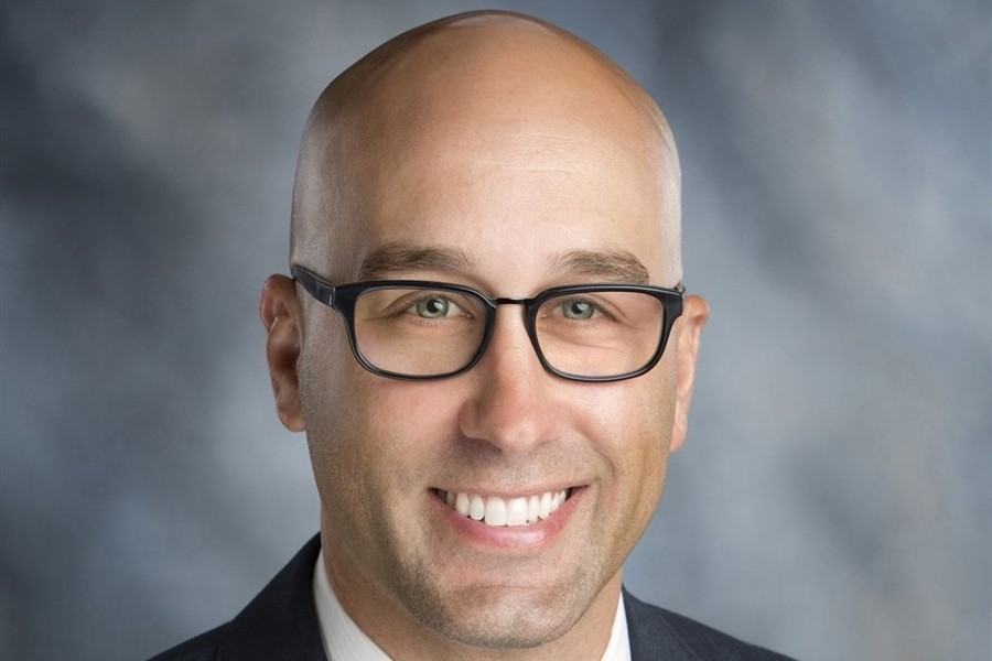Photo of a man who is bald, smiling and wearing glasses and a suit and tie. 