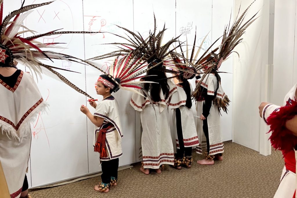 A picture of a group of children in white with feather headdresses drawing on a wall. 