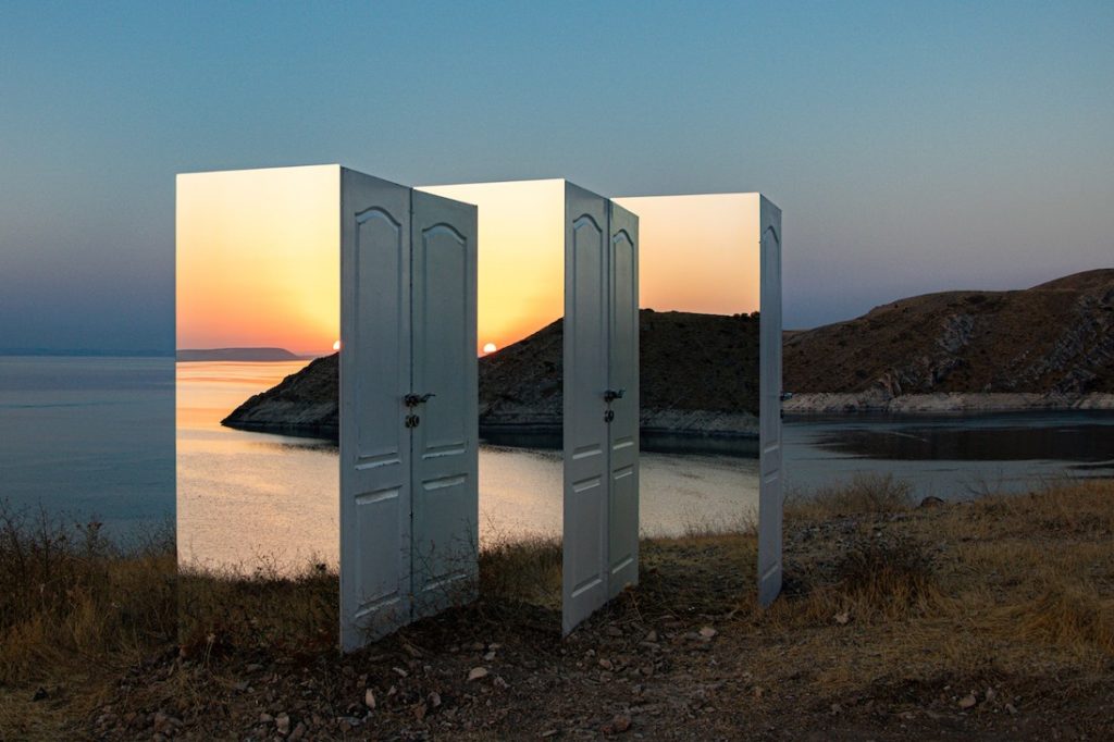 a series of three mirror panels combined with a series of three white doors at the edge of a large body of water with a large hill in the background. 