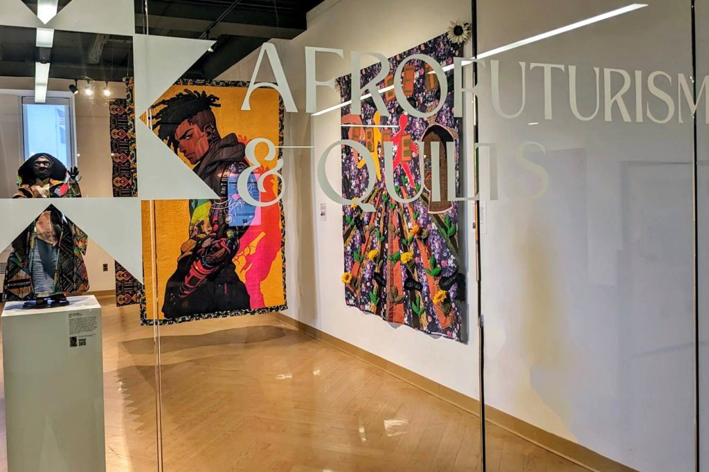 Glass wall that says: "Afrofuturism & Quilts and you can see colorful quilts through the glass. 