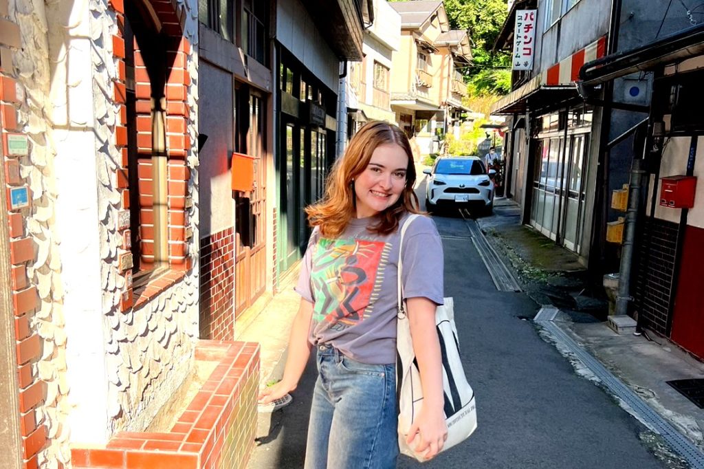 Woman wearing a T-shirt with a beige canvas bag hanging over her shoulder, standing on a narrow street in Japan. 