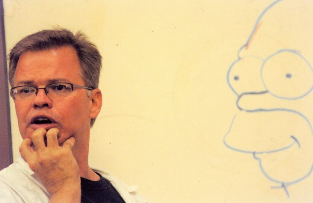 A picture of a man with short hair and glasses standing in front of a drawing.