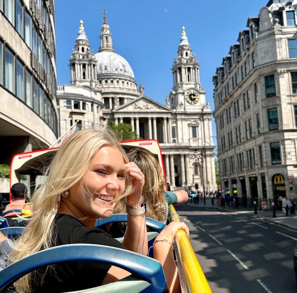 Woman riding in an open air touring vehicle with the wind blowing through her long blonde hair as she raising her hand to move the hair from her face. The vehicle is driving through the streets of London and in the background is a large ornate building with a clock on it. 