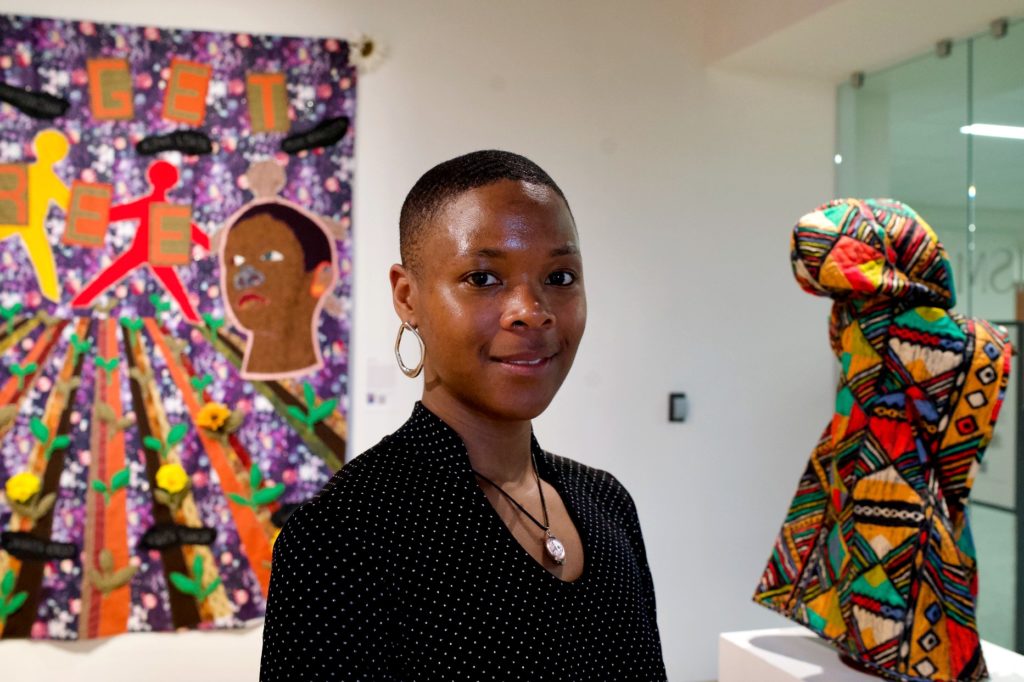 A Black person with a quilt hanging on the wall in the background.