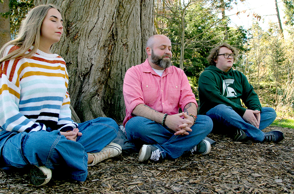 Three people sitting with their legs crossed and eyes closed outside near a tree