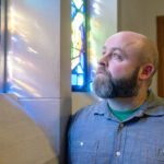 A man with a beard looks at a stained-glass window that has sunlight shining through it.