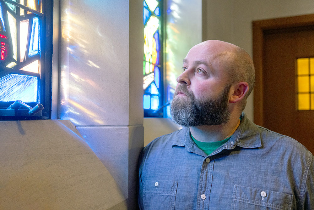 A man with a beard looks at a stained-glass window that has sunlight shining through it.