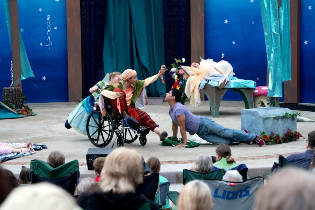 A picture of a play featuring a person in a wheelchair with a man laying on the ground acting together on stage; there is a blue background and the audience can be seen in the foreground. 