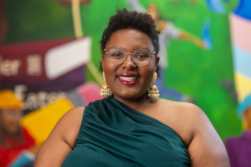 A picture of a person in a green dress wearing glasses and golden earrings in front of a a colorful background.