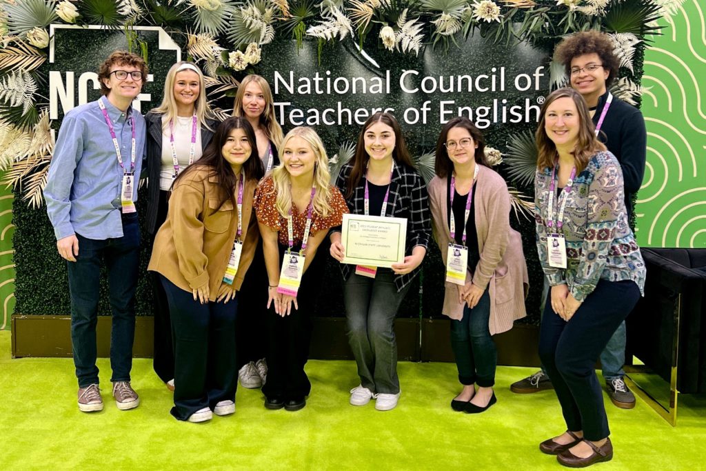 Group of nine people poising for a photo with the person in the center holding a certificate. They are standing in front of a sign that says: "National Council of Teachers of English."