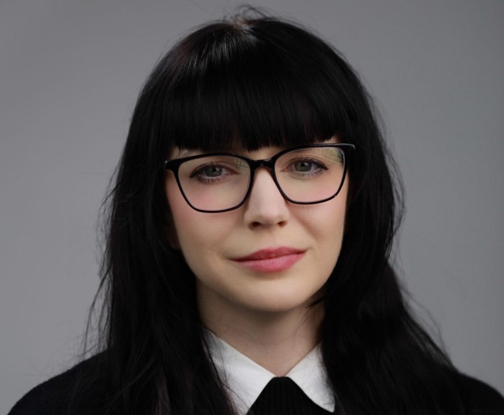 Headshot photo of a woman with long black hair, pale skin and black-rimmed glasses. 