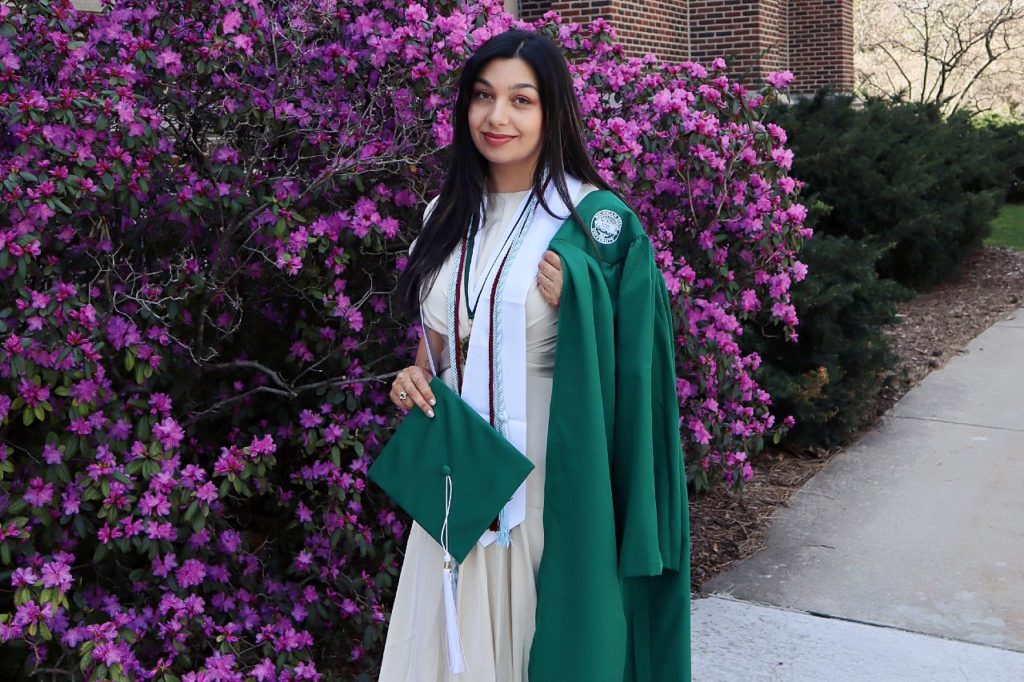 A picture of a person wearing a white dress with a dark green graduation gown in front of some purple bushes.
