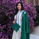 A picture of a person wearing a white dress with a dark green graduation gown in front of some purple bushes.