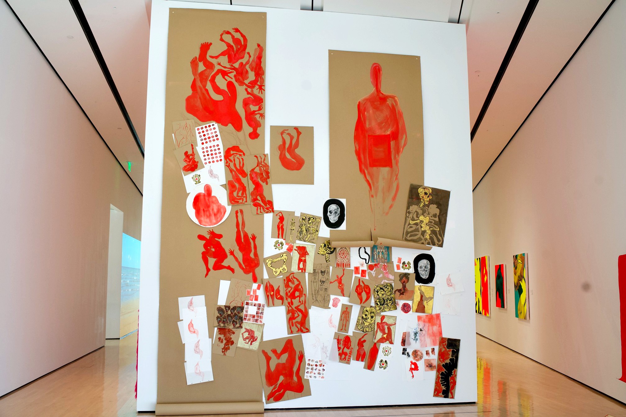 A photo of a painting in an art gallery. It is a collage of red and brown and gold art on a white wall.