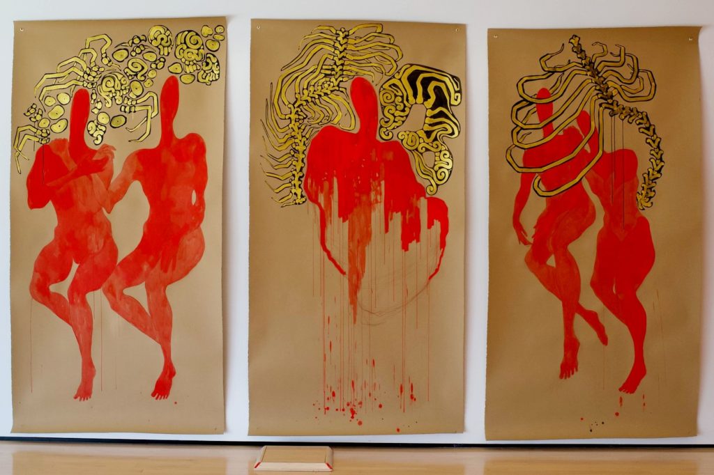 A photo of a painting in an art gallery. It is a series of three red paintings with gold accents on a white wall. 
