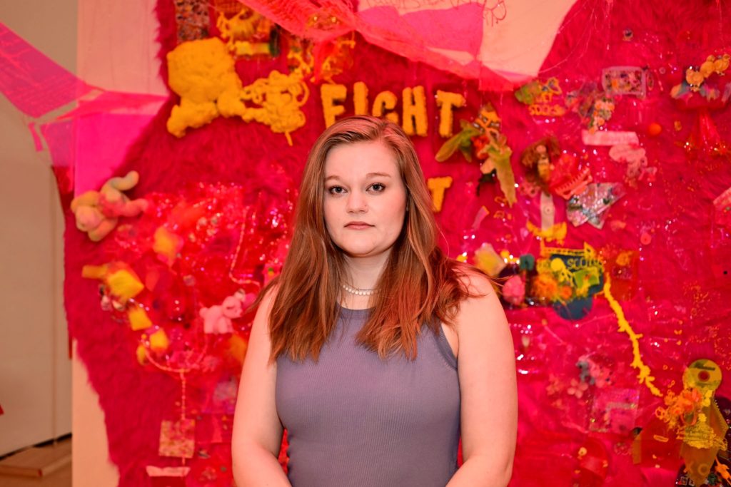 A picture of a woman with blonde hair and blue eyes standing in front of a red/hot pink wall with different toys and bits of yellow material attached to it.